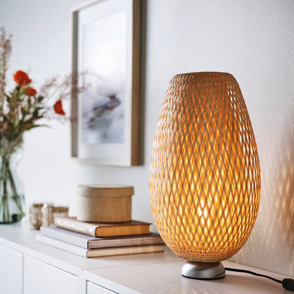Käsitööna valminud BÖJA bambuslamp kiirgab sooja valgust, seistes kõrgel raamatute ja lillekaunistuste lähedal.