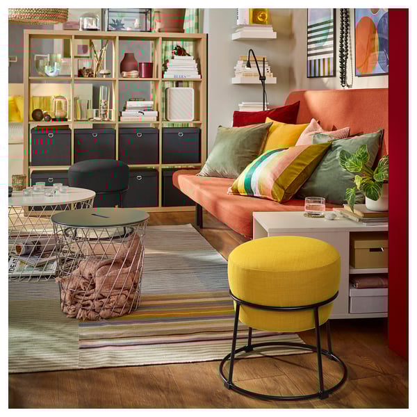 Elav elutuba: oranž diivan värviliste patjadega, kollane taburet, triibuline vaip ja kaunistusega puidust riiulid.