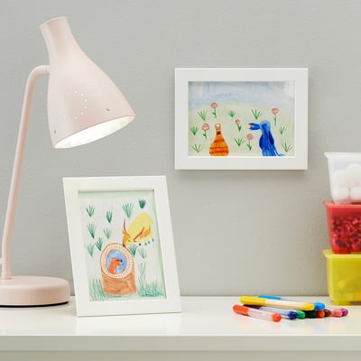 Valge kirjutuslaud, millel on raamitud värviline lastejoonistus. Selle kõrval markerid, lamp ja kollane anum pom-pomidega.