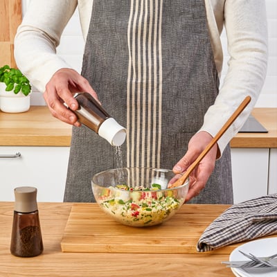 Inimene, kes maitsestab salatit pipraveskiga puidust laual. Läbipaistev klaaskauss ja maitseainepurgid nähtavad.