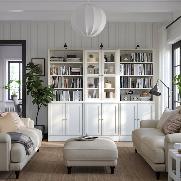 Elegantne elutuba, kus on HAVSTA valge männipuidust raamaturiiulid, diivan, ottoman, lamp, suur aken ja taimed.