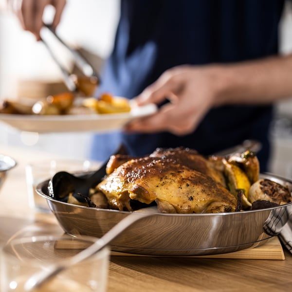 Metallist pannil röstitud kana istub puidust laual, valmis serveerimisriistadega nikerdamiseks.
