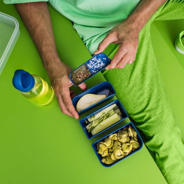 Inimene hoiab käes sinist IKEA 365+ pudelit suupistetega lahtrites rohelisel pinnal.