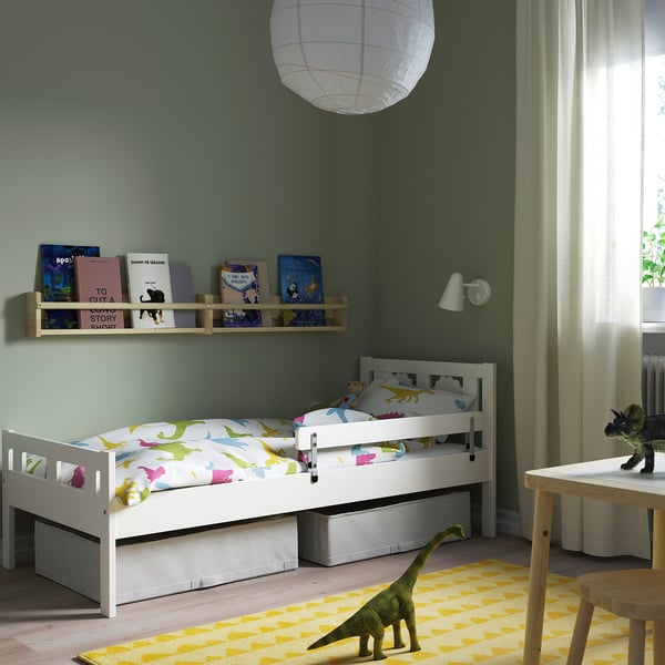 Lastetuba valge voodi, dinosauruste voodipesu, laua, kollase vaiba, raamaturiiuli ja lampidega.