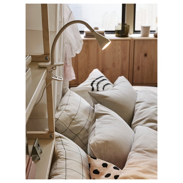 Voodipeatsi külge kinnitatud minimalistlik valge NÄVLINGE lamp, mis valgustab voodit valgete ja mustriliste patjadega.