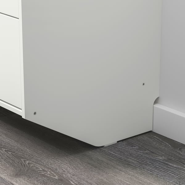 Valge RACKNÄS kummut nurgas, elegantne disain, seinale kinnitatav, tume puitpõrand.