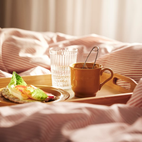 Hommikusöögikandik voodil koos SANDSKÄDDA ookerkruusi, klaasi ja taldrikuga, mis sisaldab võileiba, saiakesi ja puuvilju, rõhutades hubast maalähedast disaini.