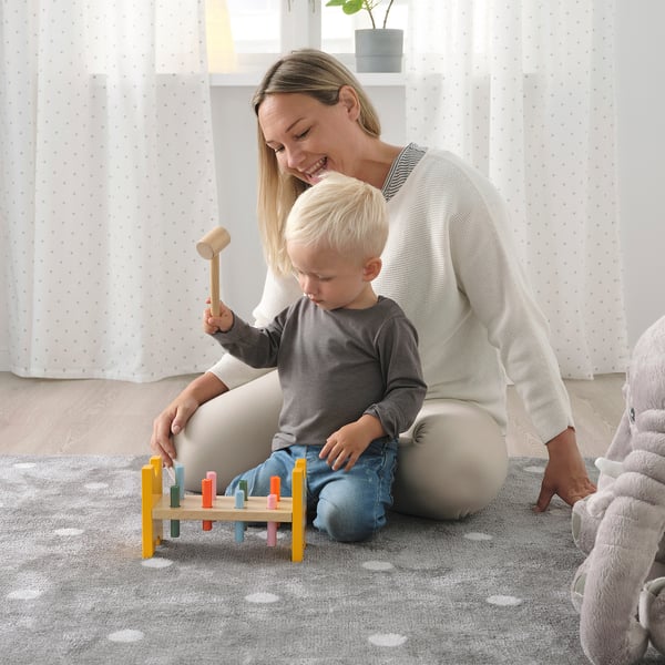 Fotol mängivad naine ja laps puidust klotsimänguasjaga vaibal, kus laps lööb haamriga plokki värvilisi pistikuid.