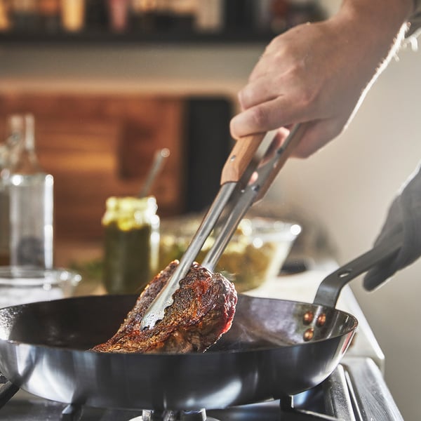 Inimene küpsetab tangidega praadi pliidil mustal süsinikterasest pannil. Pann on maitsestatud, tagades mittenakkuva küpsetamise.