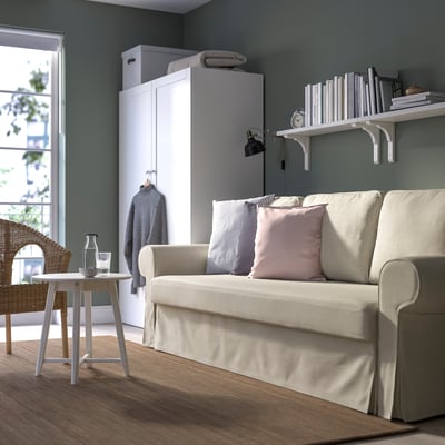 Moodne elutuba VRETSTORP beeži diivanvoodi, valgete riiulite, vitstest tooli ja kappiga. Diivanil on roosad ja triibulised padjad.