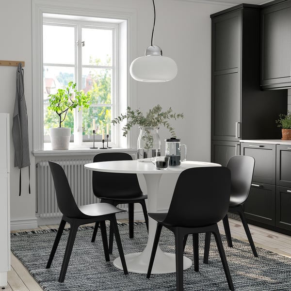 Round dining table with 4 black chairs, pendant light, and grey cabinets on a rug by a window.