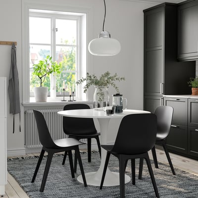 Round dining table with 4 black chairs, pendant light, and grey cabinets on a rug by a window.