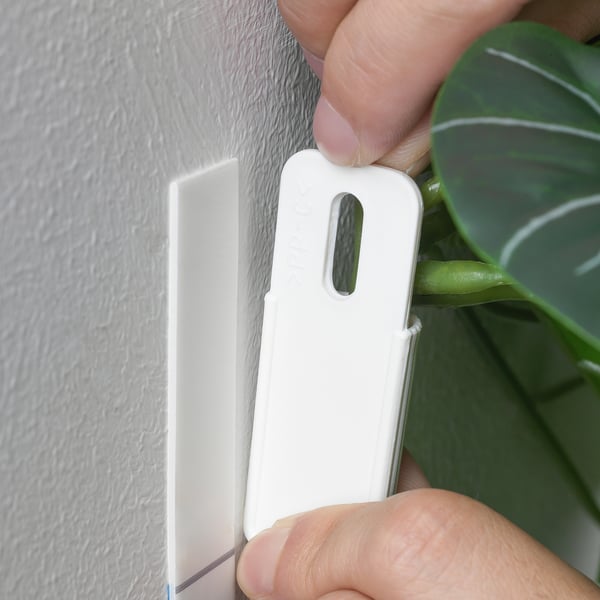 A person secures a white plant hanger on a wall, fixing an artificial plant in place.