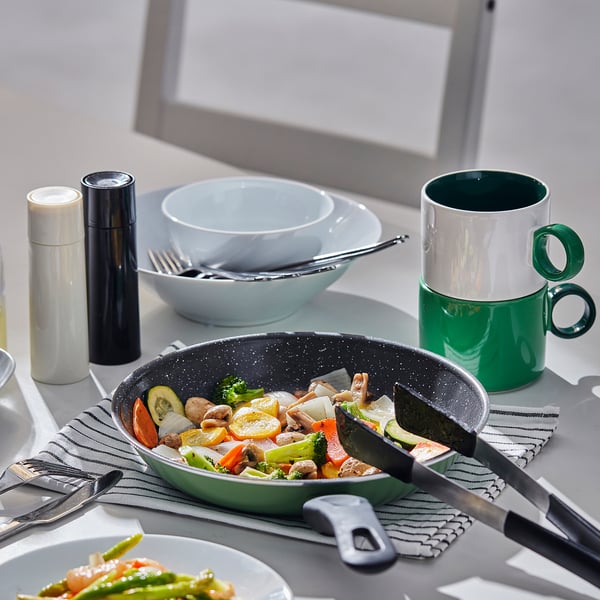 Kitchen scene: stir fry in green non-stick pan on stove, beside bowls, mugs, utensils on counter.