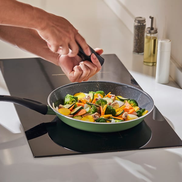 A hand sprinkles seasoning on a colourful stir-fry in a green frying pan on a black stovetop. The pan has a black handle.