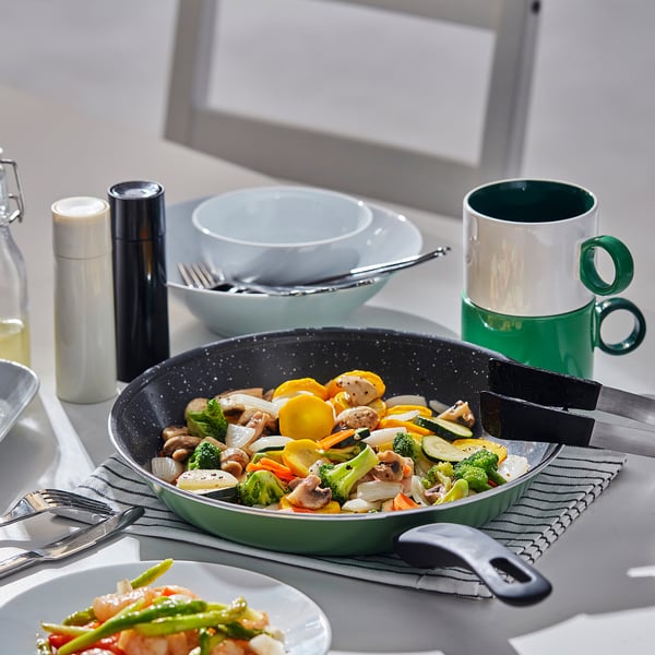Table set with a green frying pan containing stir-fried vegetables, bowls, utensils, and a green mug beside a laptop.