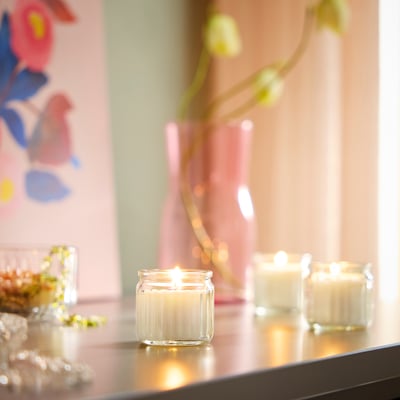 Three HÖKGÖK candles glowing on table, next to flowers and art, emitting calming vanilla scent.