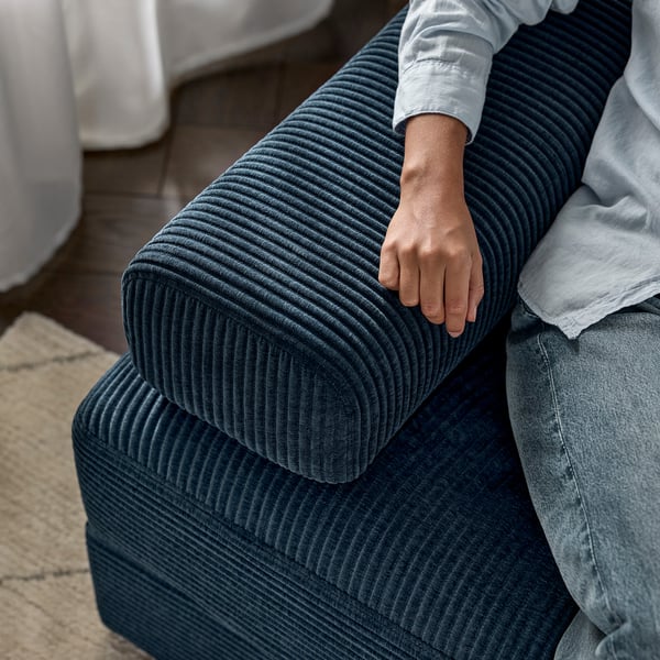 A person sitting on a blue JÄTTEBO modular sofa, resting their arm on a cushioned headrest. The sofa comfort foam and pocket springs for support.