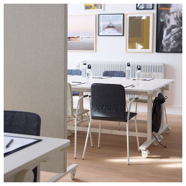 Displays a modern office space with white tables, grey chairs, and a portable acoustic screen. Decorative art fills the bright, well-lit room.