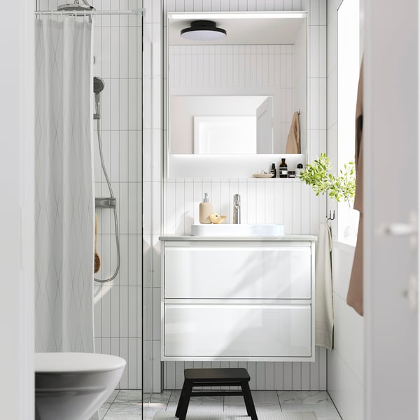 Bany modern: lavabo d'armari blanc, de paret, tamboret negre, dutxa de vidre, tovalloles, plantes.