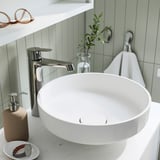 Un lavabo de bany modern amb aixeta cromada elegant, dispensador de sabó beix i petita planta en un gerro transparent sobre un taulell blanc.