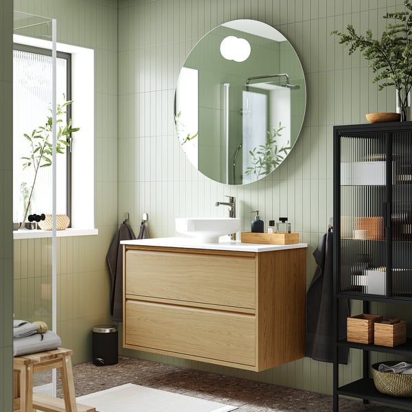 Bany modern amb tocador de fusta clara ÄNGSJÖN, taulell de marbre, lavabo rodó, mirall gran i parets de rajoles verdes.