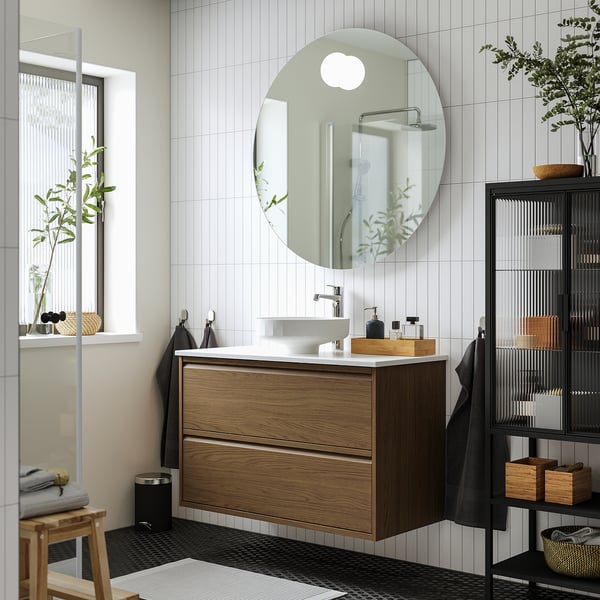 Bany modern minimalista: tocador de noguera, lavabo blanc, parets enrajolades, prestatges foscos, tovalloles, planta.