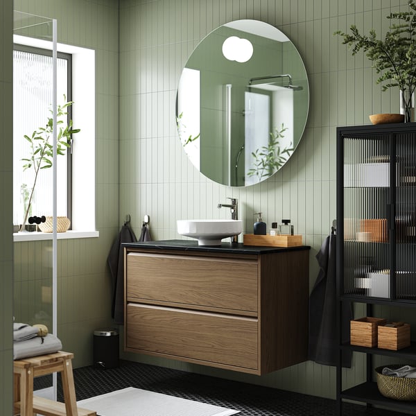 Bany amb tocador de fusta fosca, lavabo de marbre, mirall, ganxos de paret, planta, prestatgeria. Terra estampat, parets verdes, espai modern i acollidor.