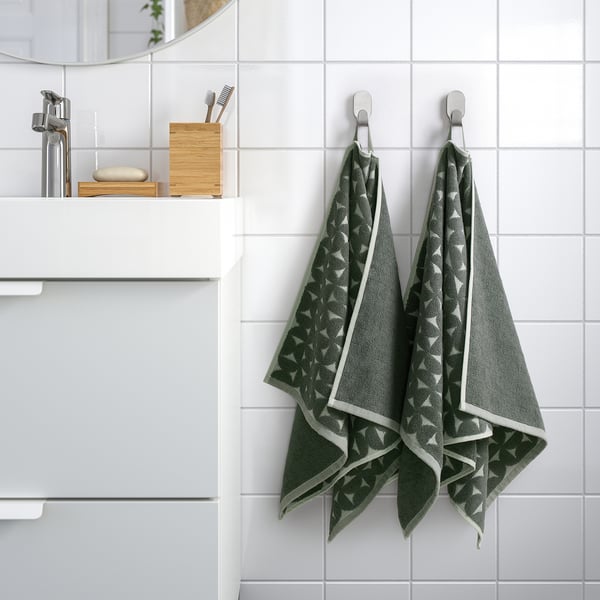 Bany modern amb dues tovalloles ÄNGSNEJLIKA amb estampats de color gris-verd penjant perfectament en ganxos, mostrant el seu disseny complex i la seva textura suau.