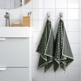 Bany modern amb dues tovalloles ÄNGSNEJLIKA amb estampats de color gris-verd penjant perfectament en ganxos, mostrant el seu disseny complex i la seva textura suau.
