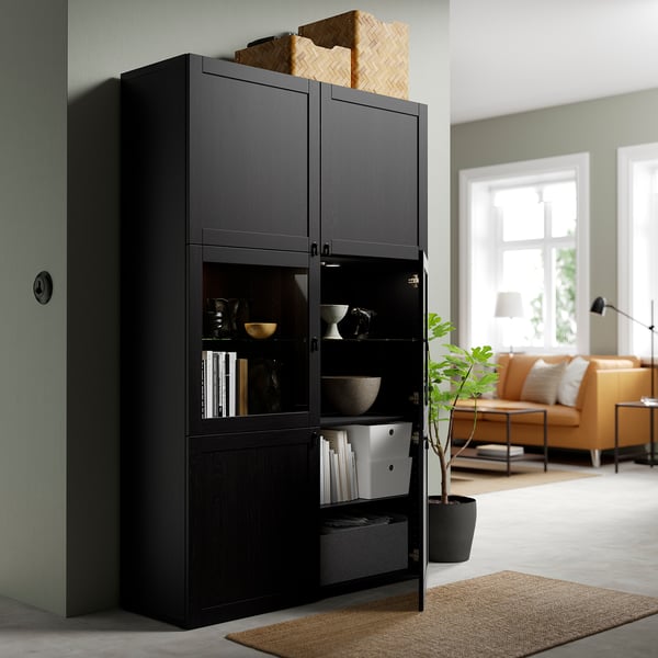 Modern armari d'emmagatzematge negre amb portes de vidre, amb decoració i llibres, situat en una acollidora sala d'estar amb sofà i planta.