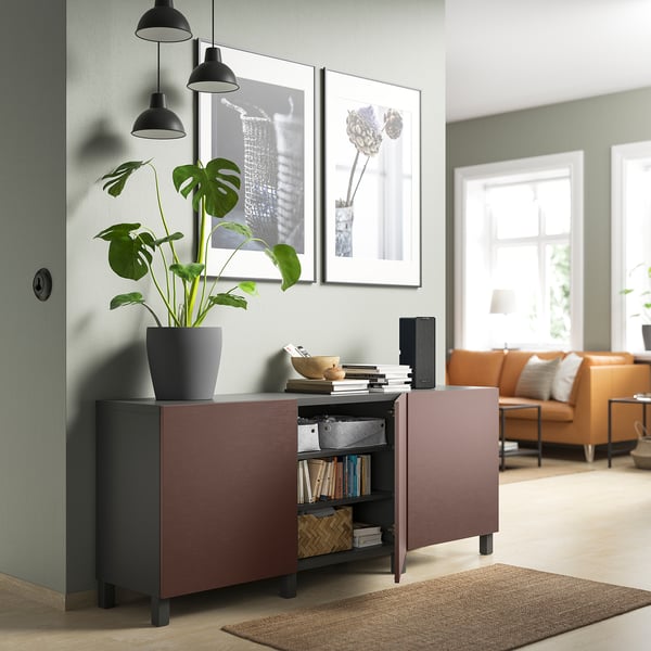 Sala d'estar moderna amb moble d'emmagatzematge BESTÅ granat, amb llibres, planta verda, llums penjants negres, sofà taronja, catifa natural i art de paret emmarcat.