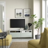 Sala d'estar moderna amb suport de TV blanc BESTÅ, TV de pantalla plana, sofà groc, planta gran en test i fotos de la natura emmarcades.