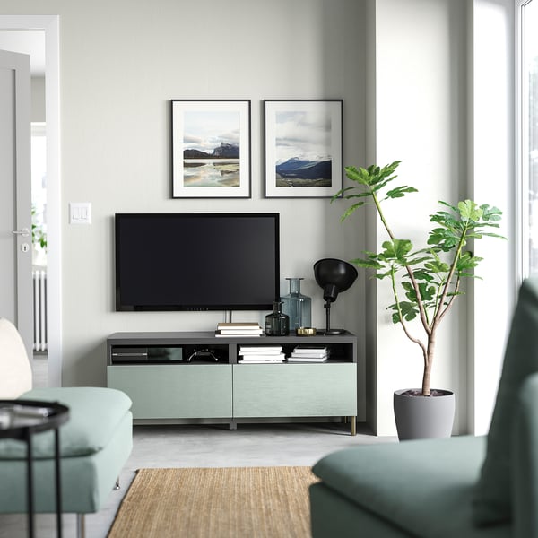 Saló amb parets grises clares, banc de televisió BESTÅ, tv, fotos, ampolles i planta prop del sofà blau.