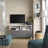 Sala d'estar moderna amb banc de televisió BESTÅ, tv, fotos, llum, llibres, gerro, cadires blaves i cistella de mantes.