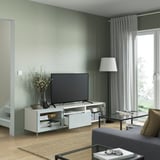 Sala d'estar moderna amb TV al suport BESTÅ, llibres, flors, comandament a distància. Gran finestral amb cortines grises, sofà acollidor, taula de centre de vidre.