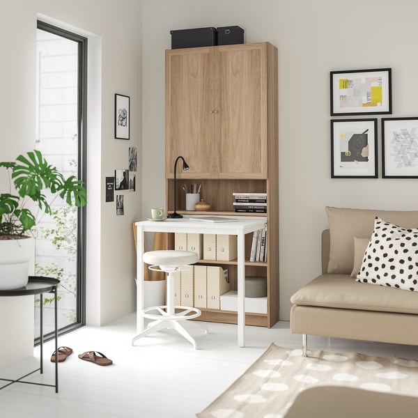 Configuració moderna de l'oficina a casa: escriptori blanc, prestatgeria BILLY de fusta clara, cadira giratòria blanca, planta en test, sofà beix amb coixí de lunars i art de paret.