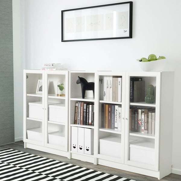 Una prestatgeria blanca BILLY amb portes de vidre, amb llibres, decoració i art. Hi ha una catifa de ratlles al davant i una imatge emmarcada penja a sobre.