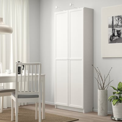 Una moderna prestatgeria blanca BILLY s'alça alta, prop d'una taula de menjador i una cadira, que ofereix un ampli espai d'emmagatzematge. El seu disseny elegant s'adapta bé a l'habitació lluminosa.