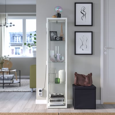 Sala d'estar moderna amb armari blanc BLÅLIDEN, prestatges decorats, marcs d'art i otomana fosca.