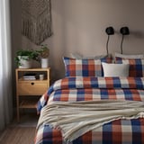 Llit amb edredó BLÅSENAP a quadres de colors, cotó suau, tapissos, tauleta de nit de fusta amb plantes i llibres.