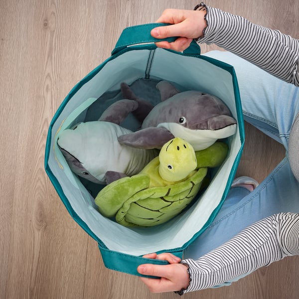 Una persona que sosté una bossa d'emmagatzematge BLÅVINGAD amb peluixos. La bossa ajuda a organitzar i transportar articles, fets de plàstic reciclat destinat a l'oceà.