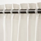 Les cortines blanques pengen amb plecs, deixant passar la llum alhora que proporcionen privacitat. Vareta metàl·lica i ganxos visibles a la part superior.