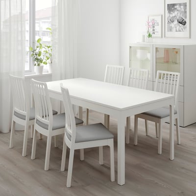 Taula de menjador blanca minimalista amb 6 cadires coixinades grises, respatllers moderns de llistons verticals, adossada a una finestra.