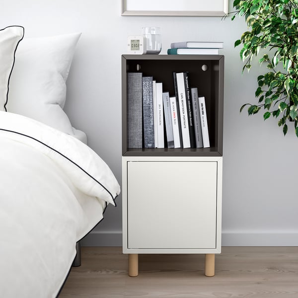 Tauleta de nit moderna amb prestatgeria EKET, llibres, rellotge i gerro. Llit proper, terra de fusta, decoració minimalista.