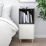 Tauleta de nit moderna amb prestatgeria EKET, llibres, rellotge i gerro. Llit proper, terra de fusta, decoració minimalista.