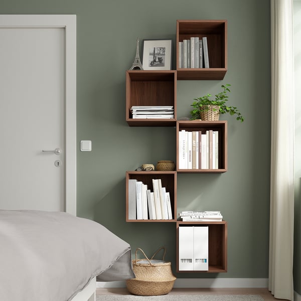 Dormitori modern amb prestatges asimètrics EKET de color marró fosc muntats a la paret, amb llibres, decoració i una planta, al costat d'un llit blanc i una porta blanca tancada.