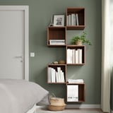 Dormitori modern amb prestatges asimètrics EKET de color marró fosc muntats a la paret, amb llibres, decoració i una planta, al costat d'un llit blanc i una porta blanca tancada.