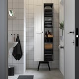 Bany modern amb armari alt blanc i prestatgeria oberta. Compacte però espaiós, conté tovalloles, ampolles i decoració. Accents negres.