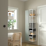 Habitació moderna amb magatzem blanc ENHET, bols, llibres i caixes. Al costat, una cadira de vímet i una finestra amb llum penjant. Parets suaus de color beix.